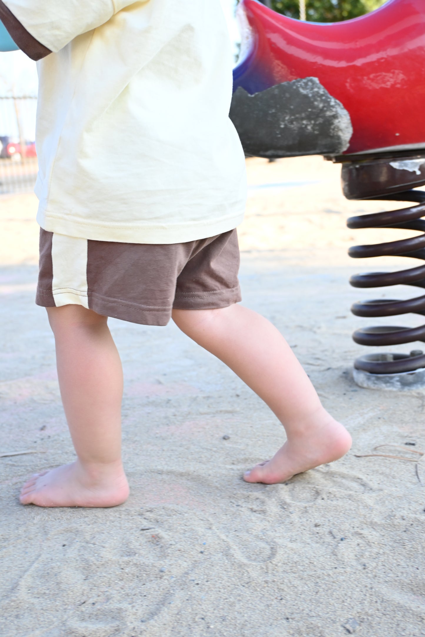 Barefoot toddler in shorts and t-shirt walking on sandy playground near spring rider toy