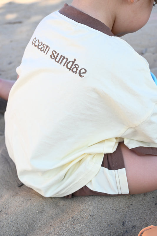 Child sitting on sandy beach wearing cream shirt with brown collar and text 'ocean sundae'