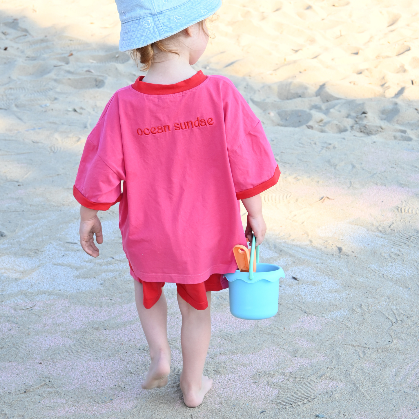 Child in pink 'ocean sundae' shirt and blue bucket hat walking barefoot on sandy beach with blue sand pail