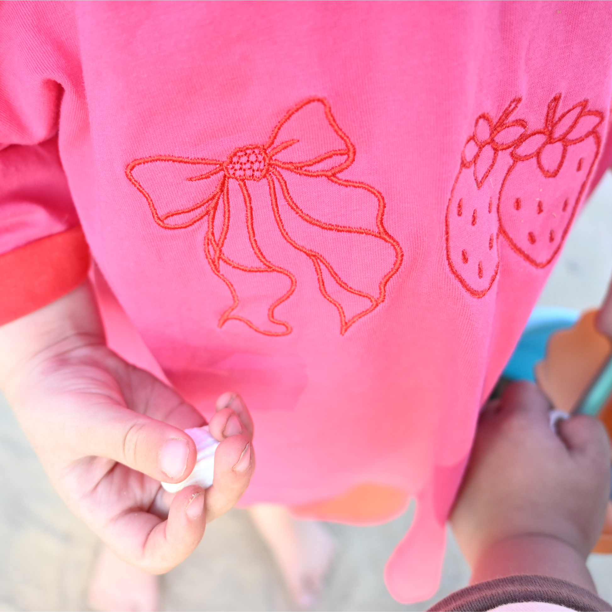 Child wearing pink shirt with embroidered bow and strawberries holding a seashell on sandy beach