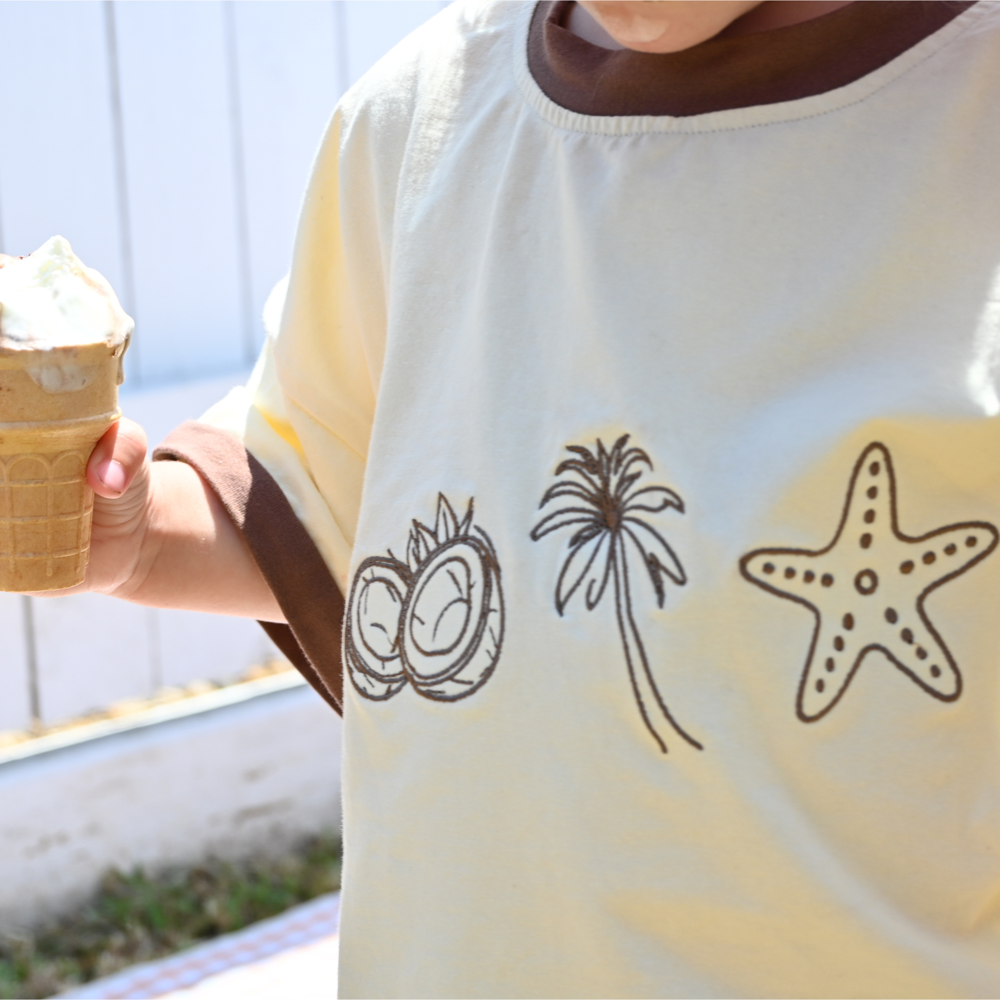 Child wearing cream shirt with coconut, palm tree, starfish designs holding melting ice cream cone