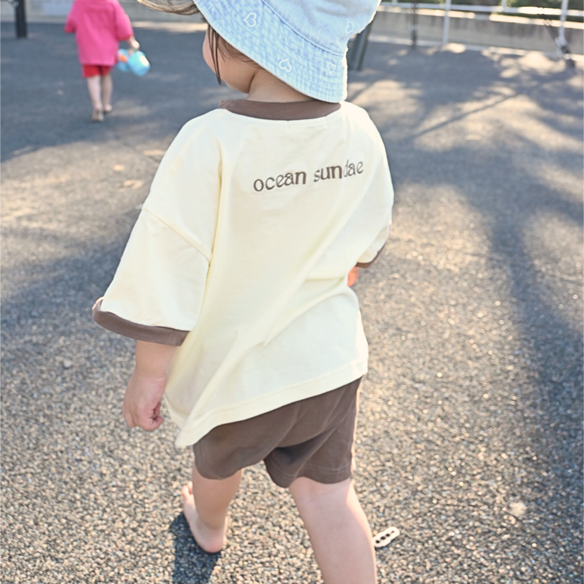 Toddler wearing a yellow and brown ocean sundae t-shirt, blue heart-patterned bucket hat, walking outdoors on sunny day