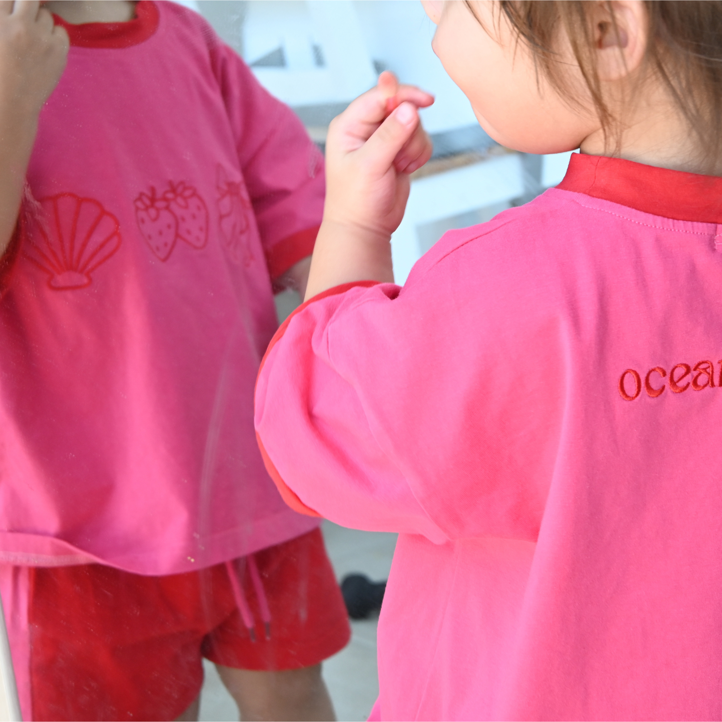 Toddler in pink and red clothing with ocean-themed prints standing in front of a mirror indoors