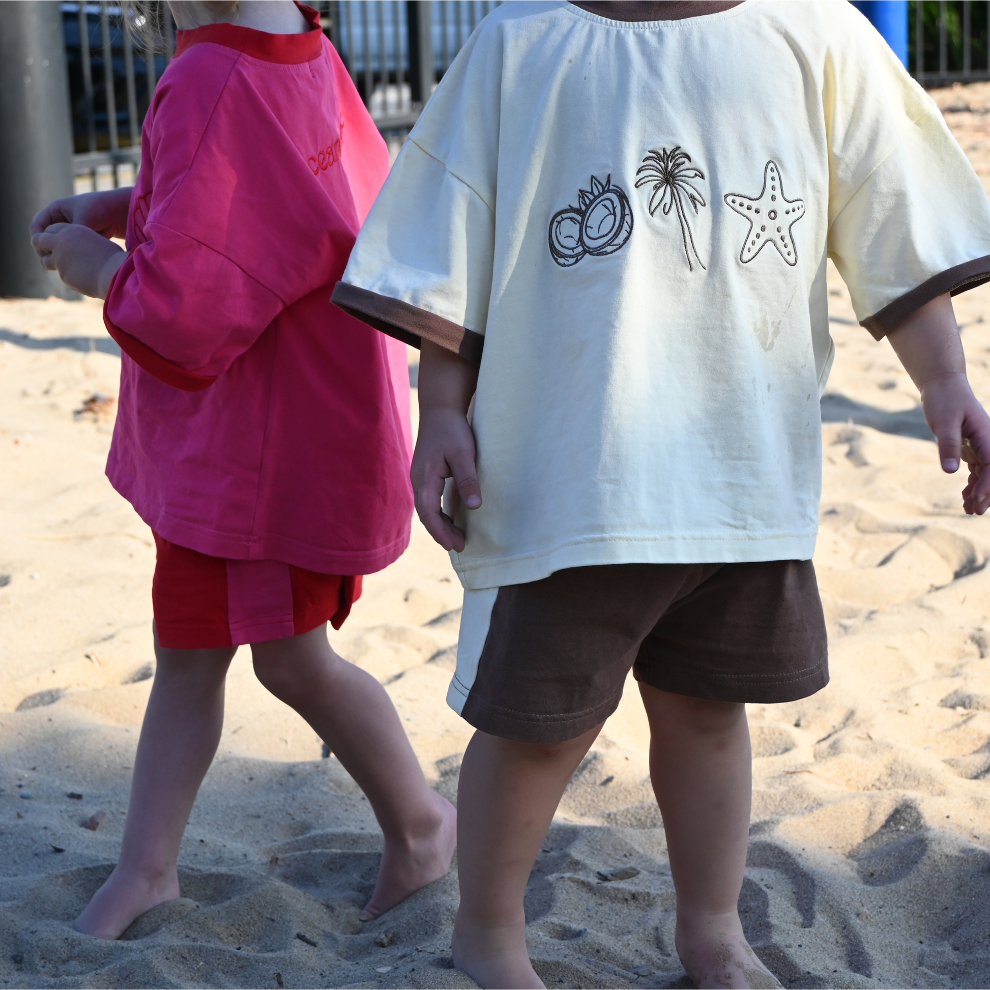 Two children on sandy beach wearing summer outfits with tropical prints and shorts