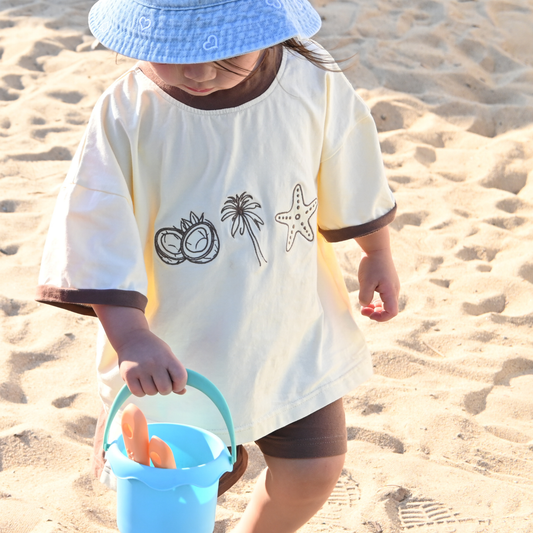 Toddler in blue hat and cream beach shirt with coconut, palm, starfish holding blue sand bucket