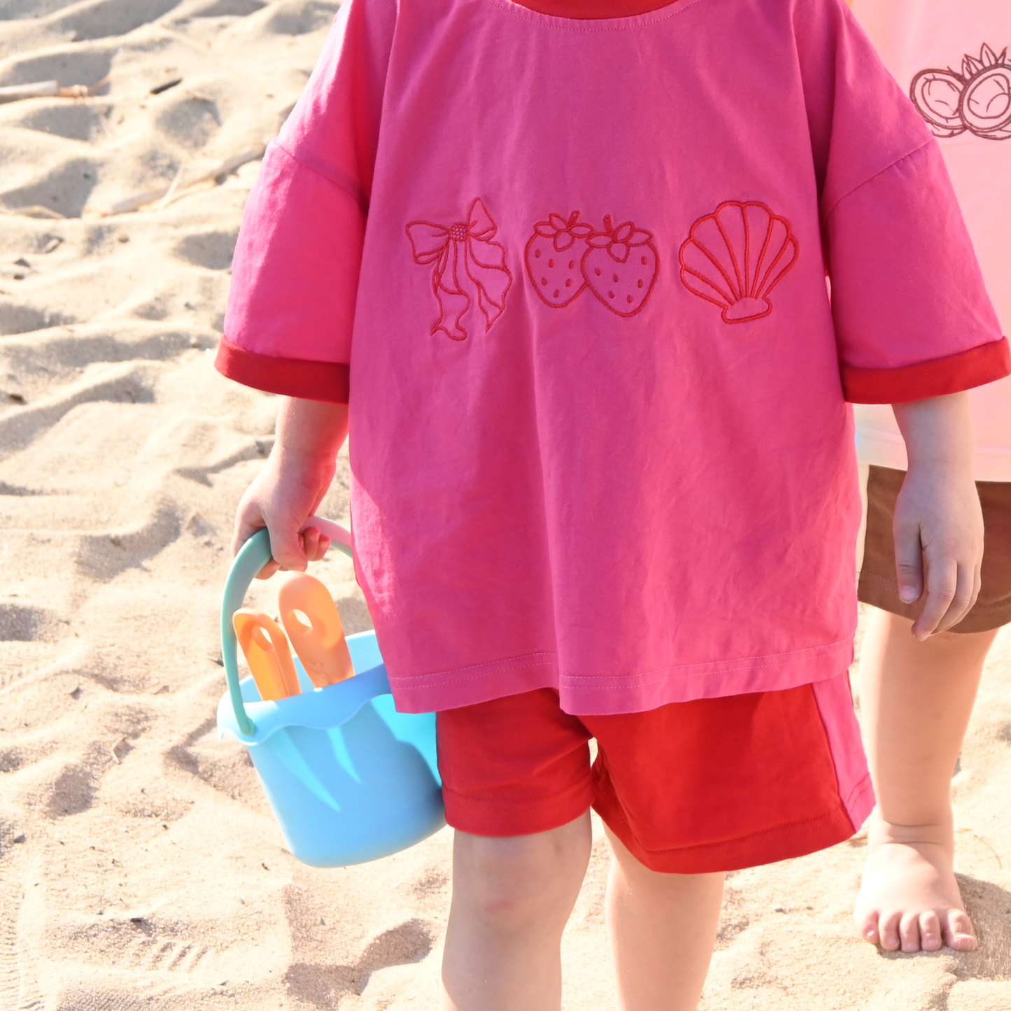 Child in pink and red outfit holding blue bucket with orange sand toys on sandy beach