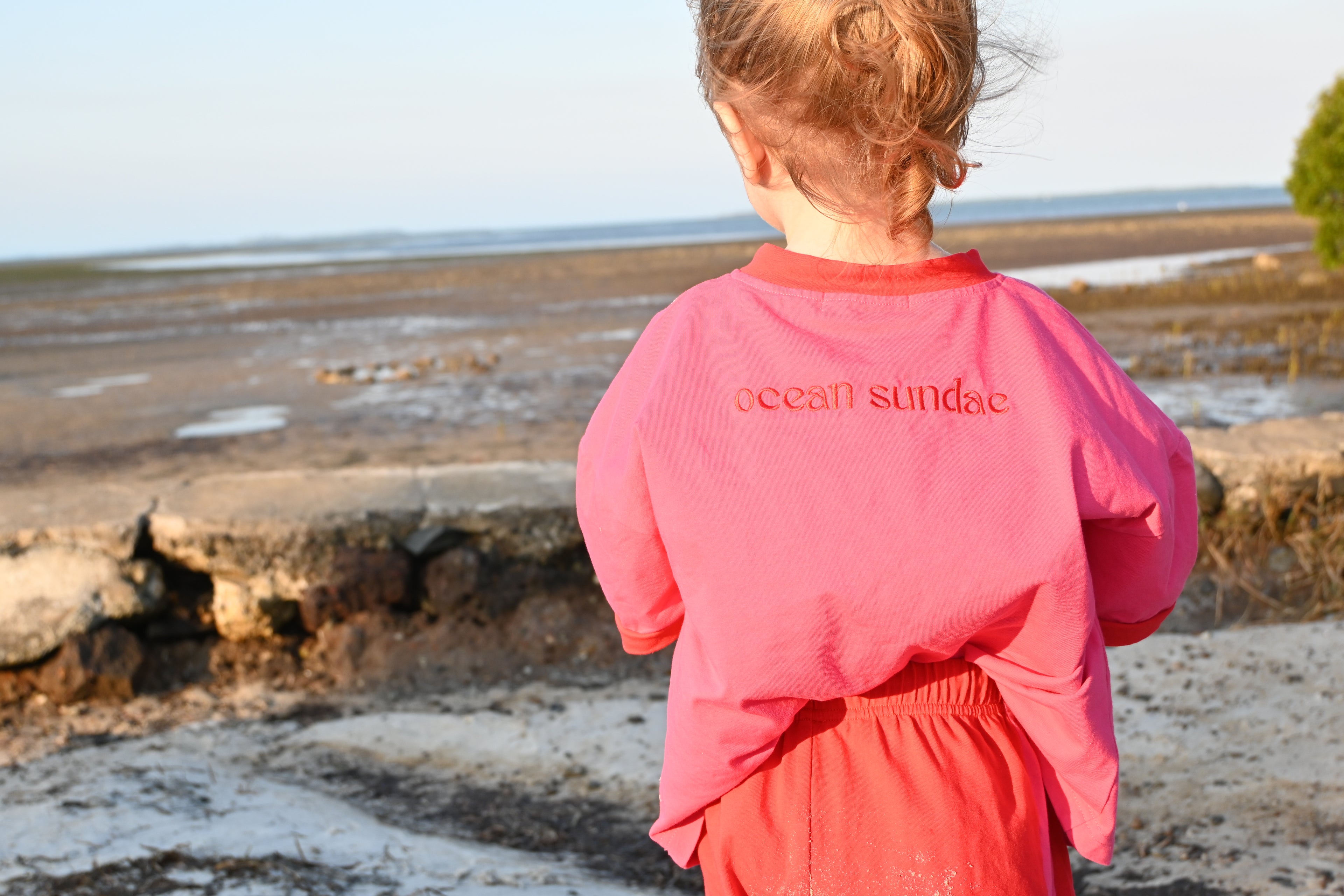 Child wearing a pink and red 'ocean sundae' shirt standing on a beach.