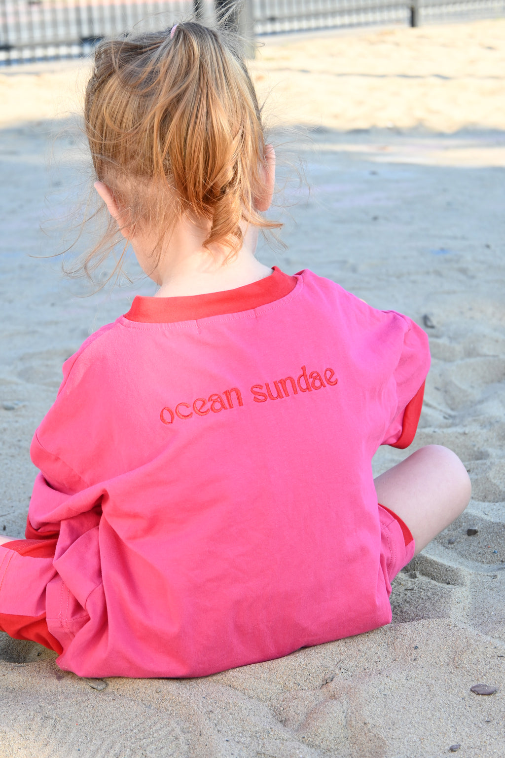 Child wearing a pink shirt with 'ocean sundae' text on a sandy surface