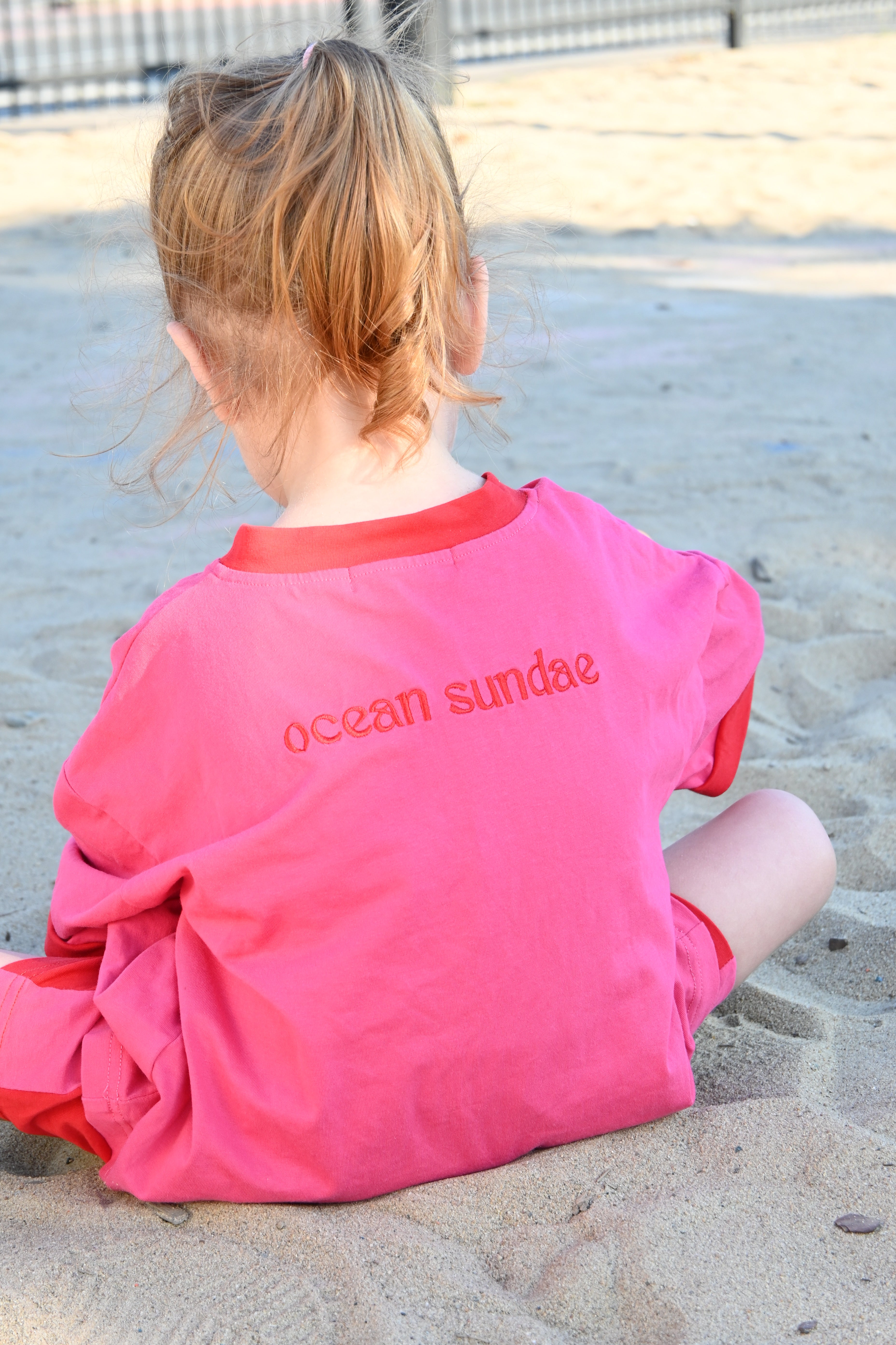 Child wearing a pink shirt with 'ocean sundae' text on a sandy surface