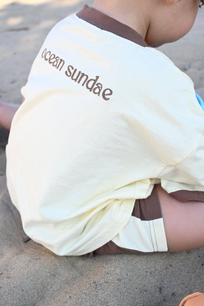 Child sitting on sandy beach wearing cream shirt with brown collar and text 'ocean sundae'