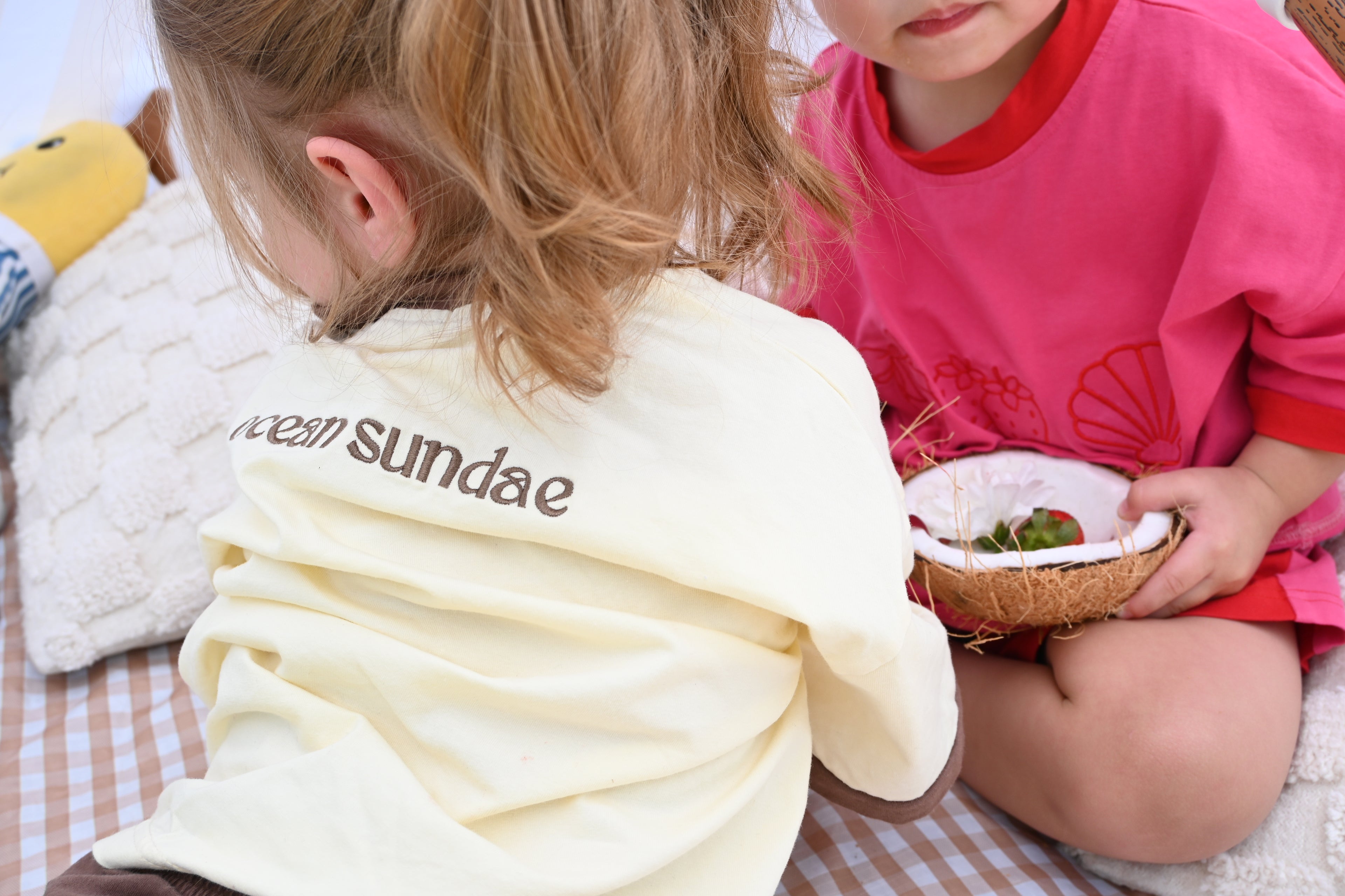 Two children sitting on a picnic rug, one wearing a beige shirt with 'ocean sundae' text, the other in a pink shirt holding a coconut with strawberries in it