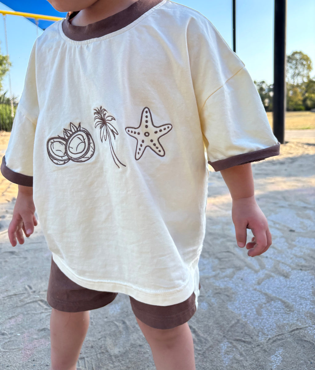 Child wearing a white shirt with brown trim and beach-themed designs outdoors.