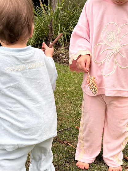 Two children outdoors, one in light blue and the other in pink outfits, holding sticks and flowers
