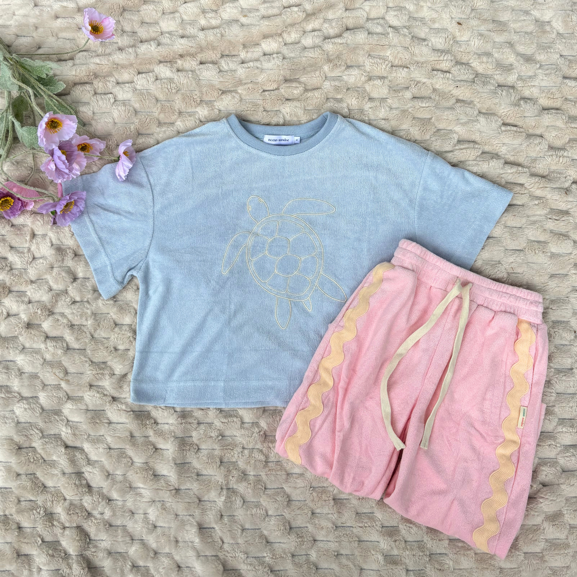 Light blue t-shirt with a turtle design and pink shorts on a woven surface with flowers.