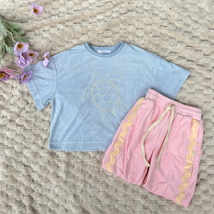Light blue t-shirt with a turtle design and pink shorts on a woven surface with flowers.